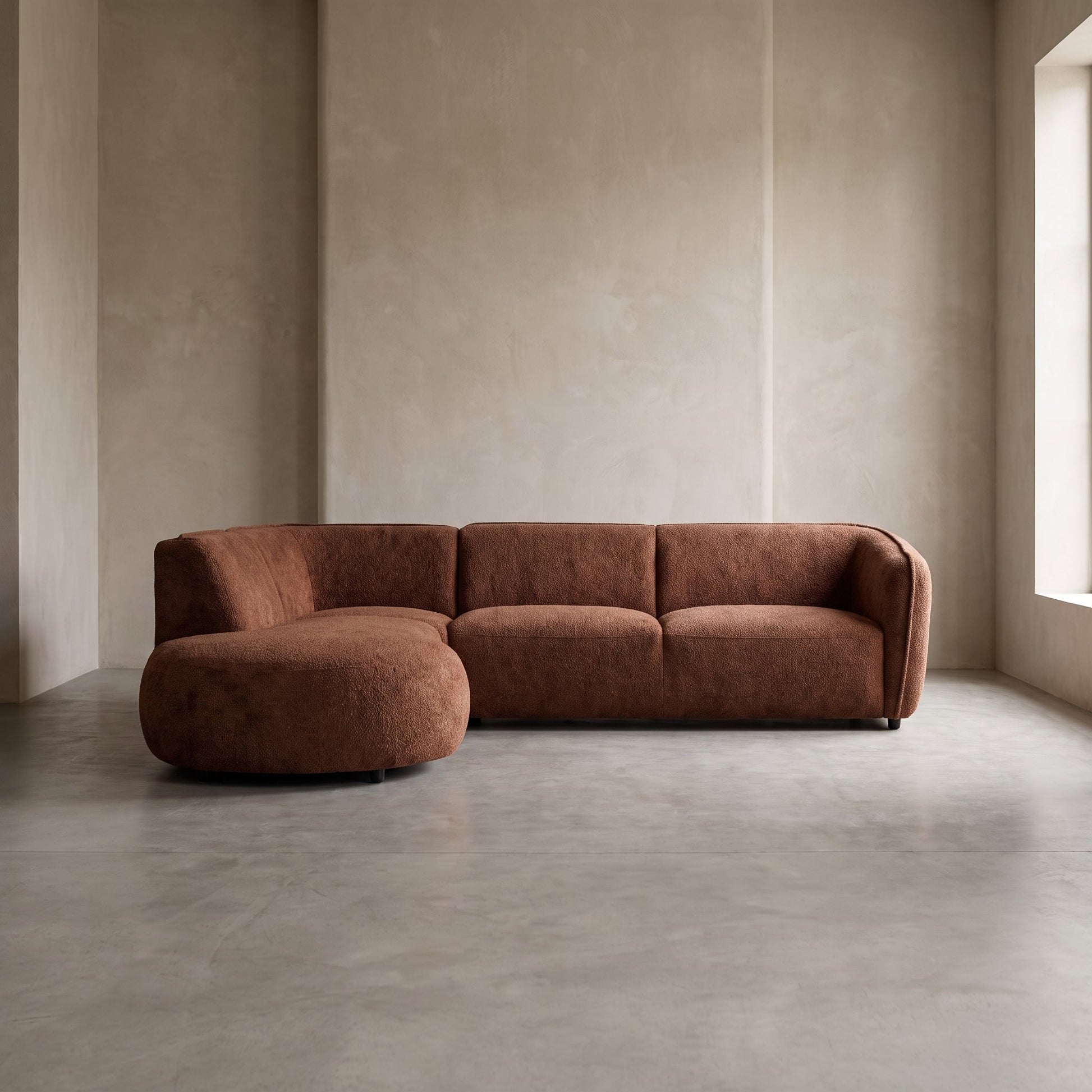 A modern curved sectional sofa in a warm rust-brown textured fabric, set in a minimalist room with concrete floors and textured beige walls.